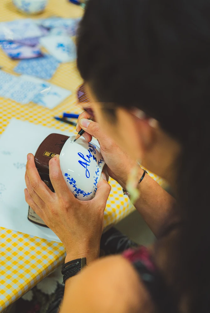 A person decorates a white ceramic bowl with blue writing and floral patterns.