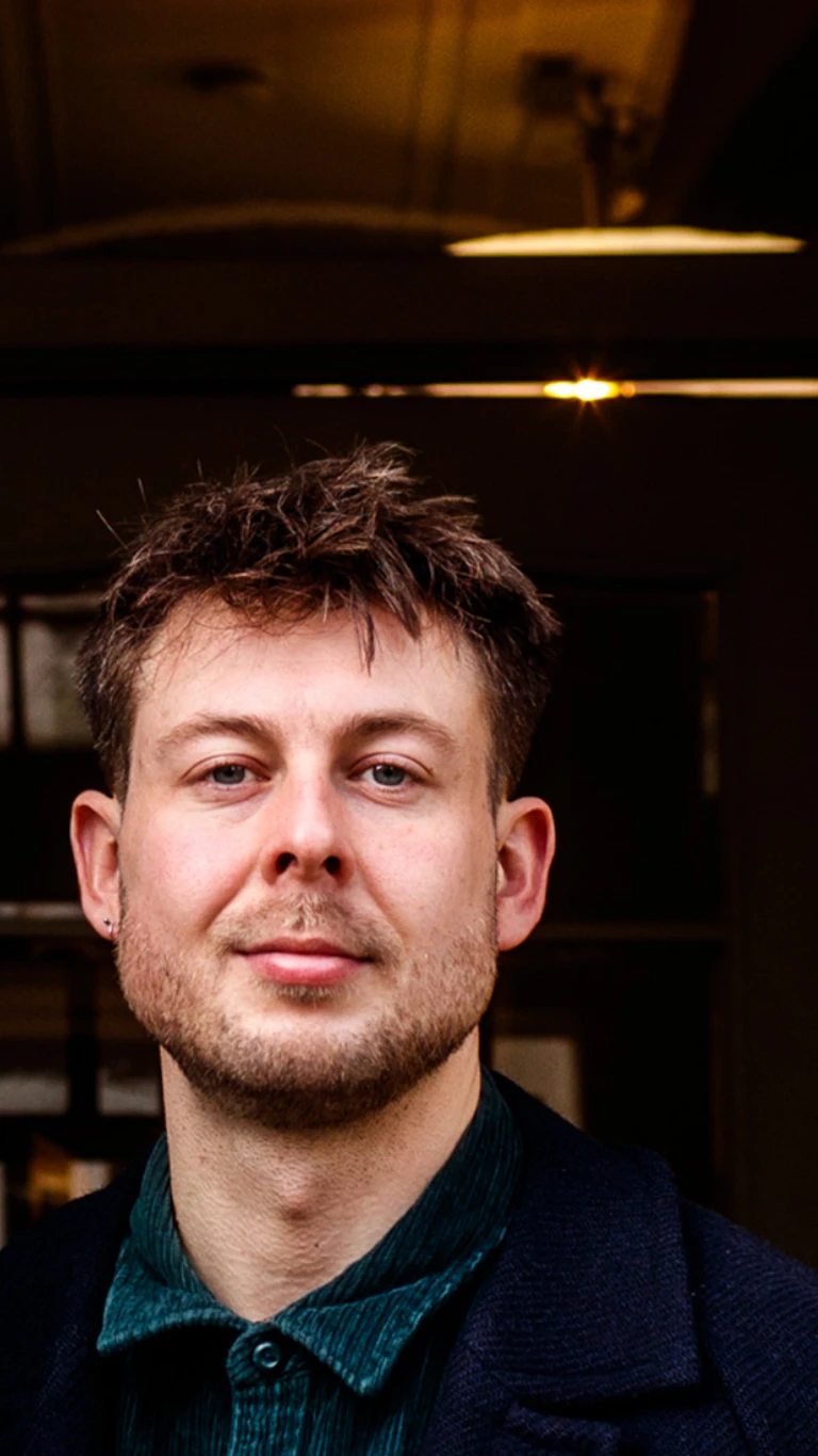 Portrait of a man with short brown hair and a beard, wearing a dark coat and green shirt indoors.