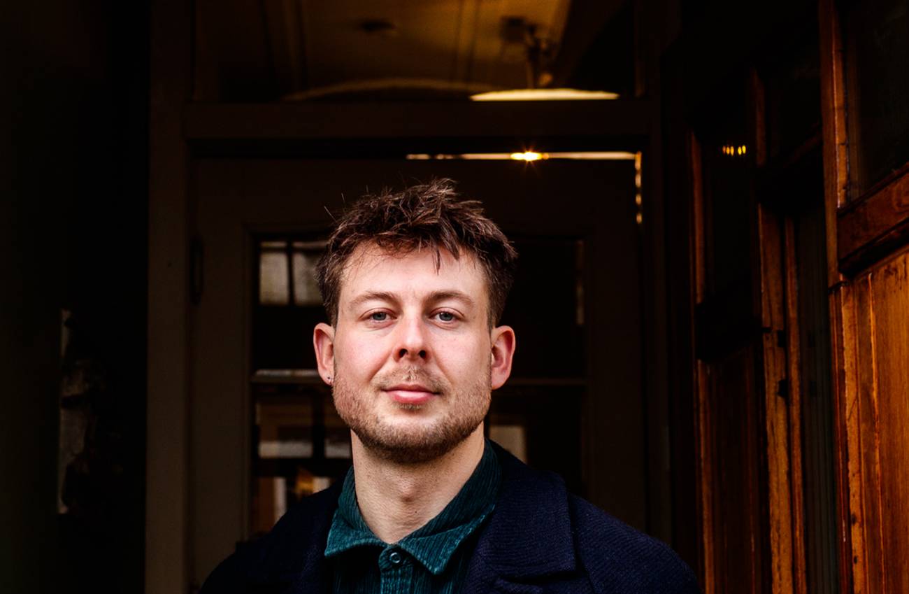 Portrait of a man with short brown hair and a beard, wearing a dark coat and green shirt indoors.