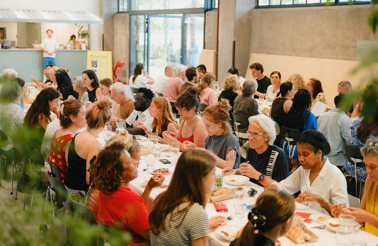 twee lange gedekte tafels waar een divers gezelschap aan zit te eten, de sfeer is zomers
