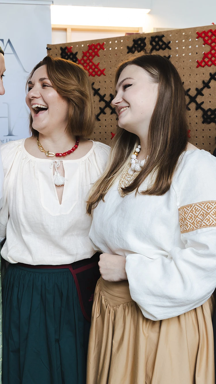 Four women in traditional attire stand smiling together. They wear white embroidered blouses.