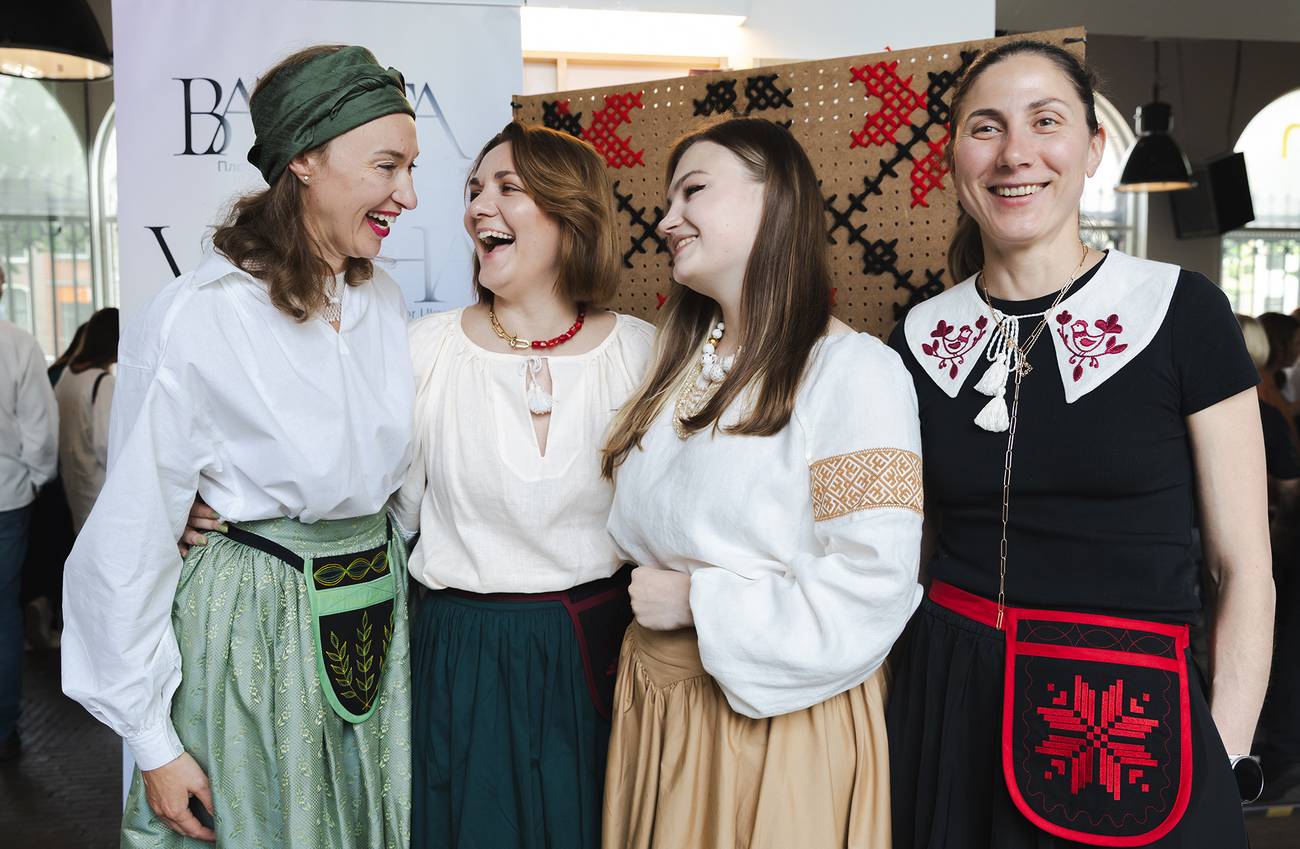 Four women in traditional attire stand smiling together. They wear white embroidered blouses.