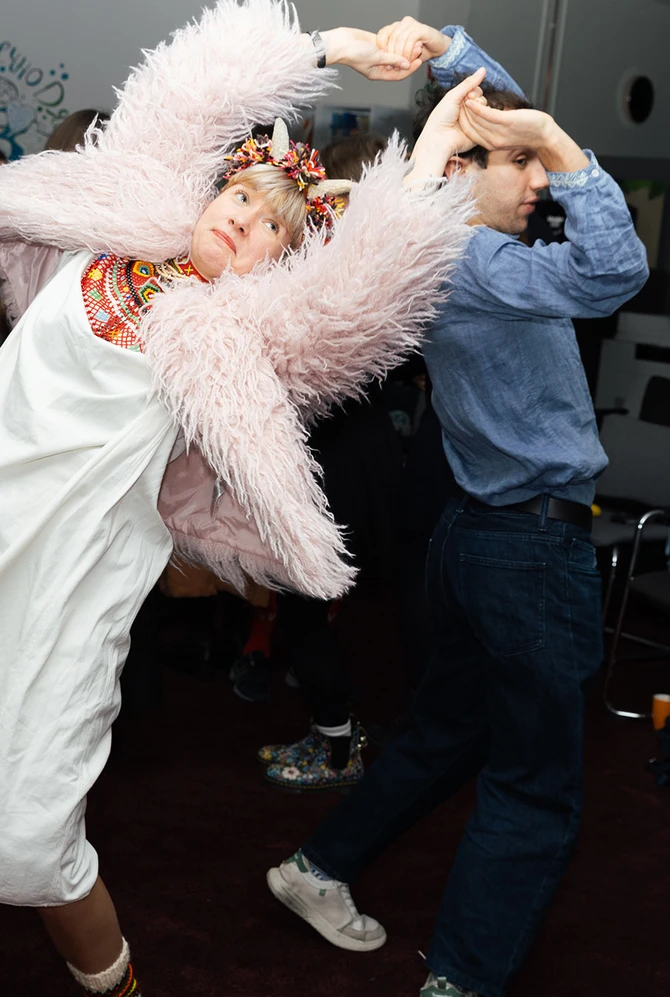 A woman in a fluffy pink coat and traditional dress dances energetically with a partner.