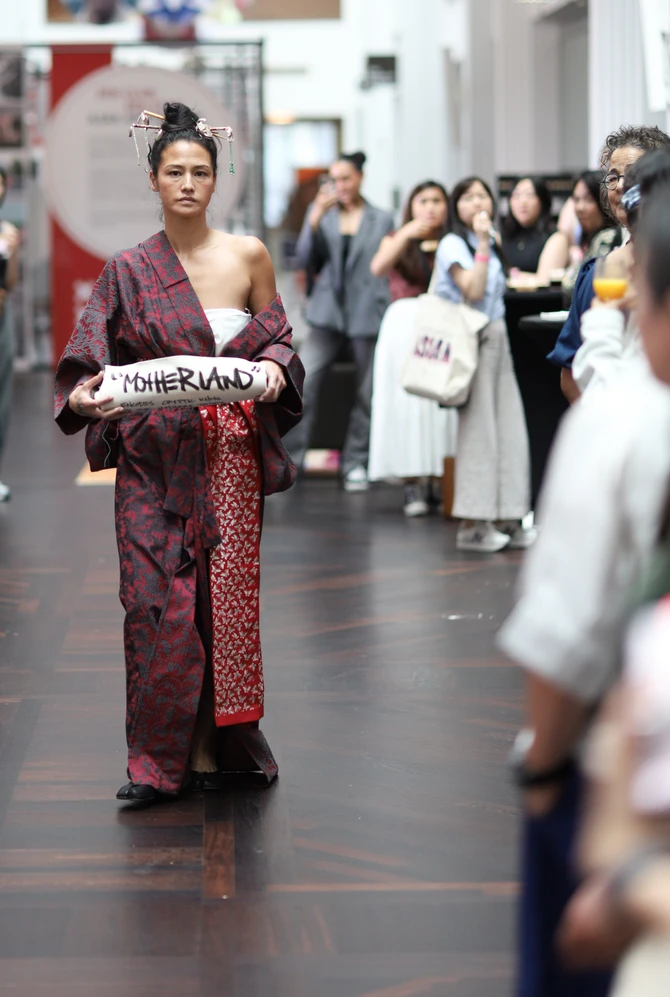 A person walks confidently down a catwalk in a red-patterned robe, holding a pillow labeled "Motherl