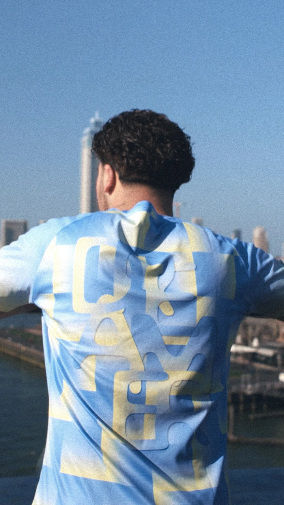 A person in a blue and yellow shirt stands on a balcony, overlooking a cityscape