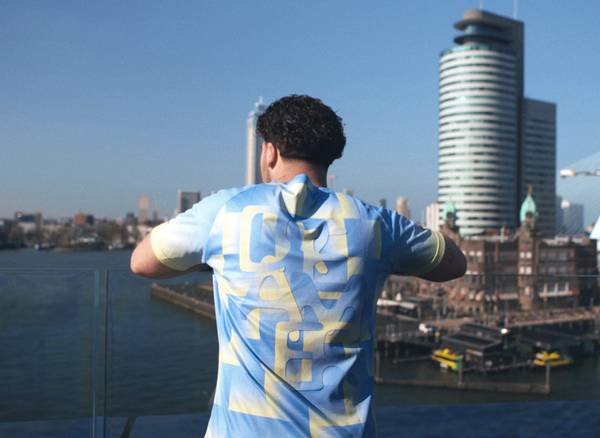 A person in a blue and yellow shirt stands on a balcony, overlooking a cityscape