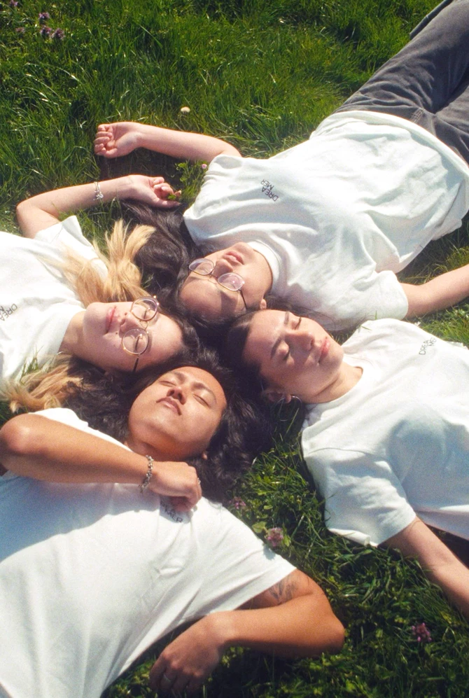 Four people in white t-shirts lie on green grass in a circle, heads together, eyes closed.