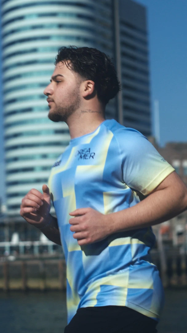 A man jogs along a waterfront in a bright, patterned shirt. Modern skyscrapers and a bridge are in