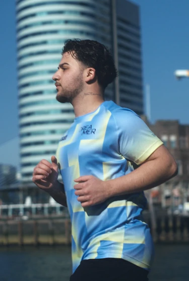 A man jogs along a waterfront in a bright, patterned shirt. Modern skyscrapers and a bridge are in