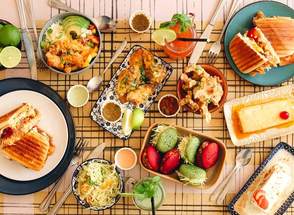 Gedekte tafel met sandwiches, salades, desserts en drankjes, kleurrijk gebak en diverse sauzen.