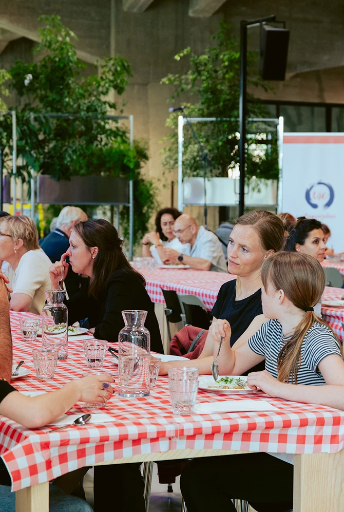 Mensen aan tafels met rood-wit geblokte tafelkleden, eten en praten in een ruimte met veel groen.