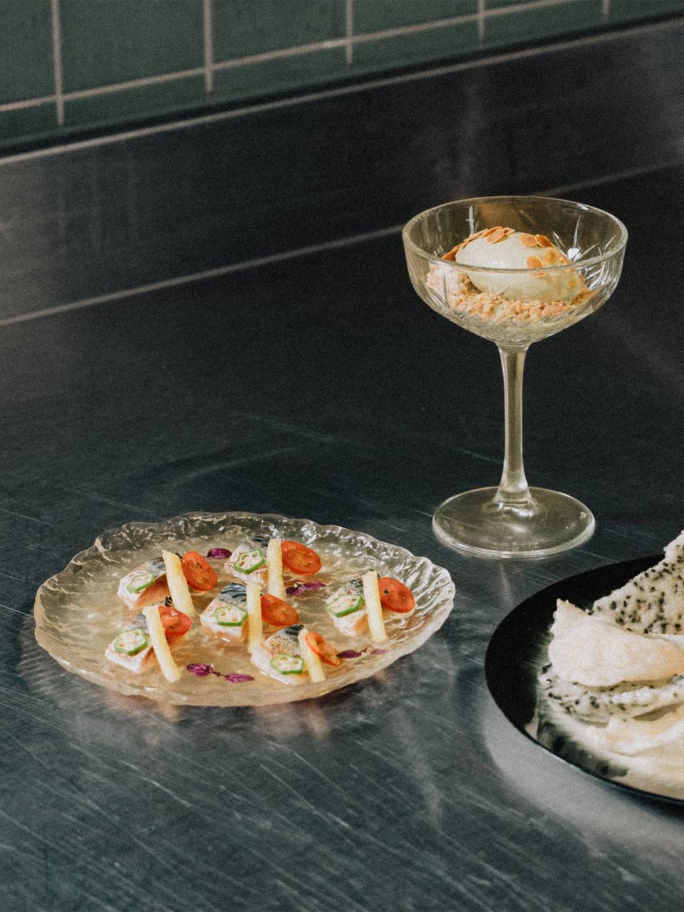 Elegant dish trio on a stainless-steel counter: mackerel, dessert in a glass, and tartar.