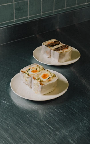 Two plates of neatly wrapped sandwiches sit on a stainless steel counter.