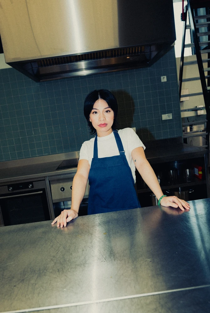 A confident woman in a blue apron leans on a stainless steel counter in an industrial kitchen.
