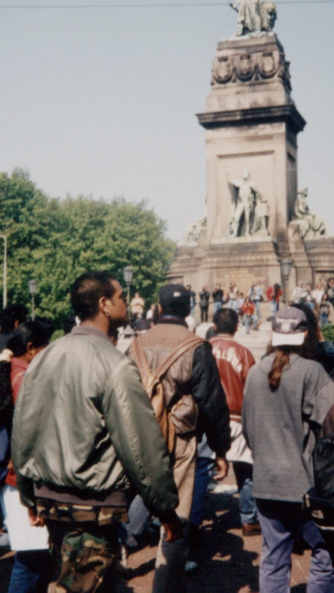 A large protest gourp walks towards a grand monument with statues, surrounded by green trees