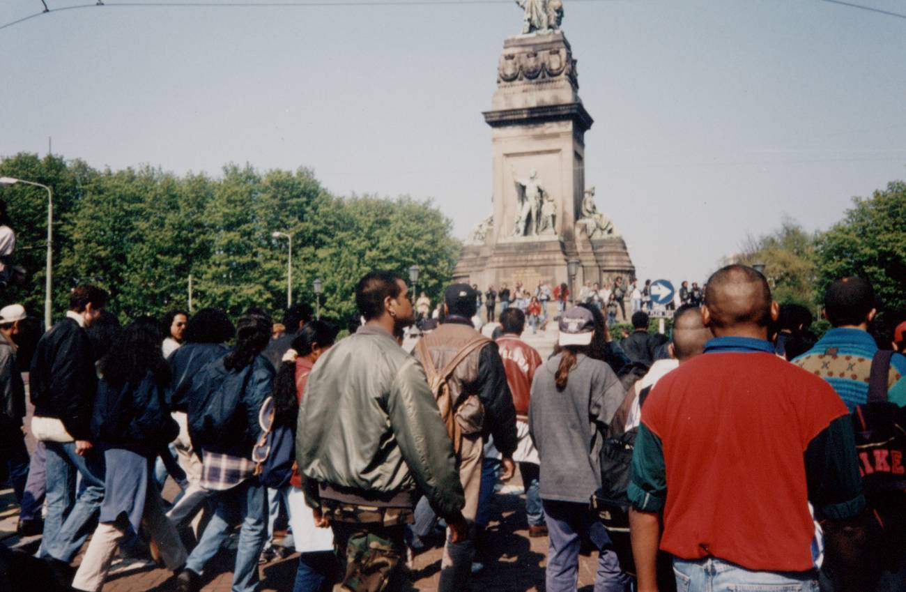 A large protest gourp walks towards a grand monument with statues, surrounded by green trees