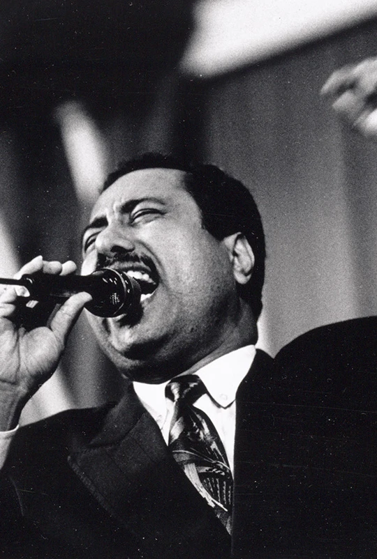 A man passionately sings into a microphone, wearing a suit and tie, with his fist raised.