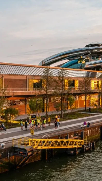 People stroll along a riverside path beside a modern industrial building at sunset.