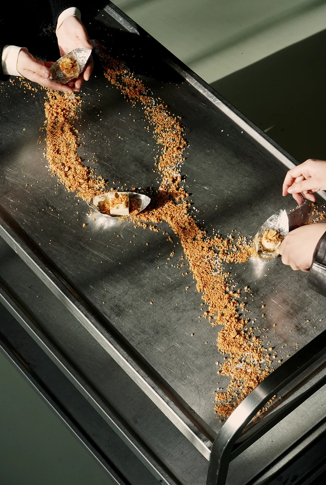 Two hands hold desserts on a metal table with a trail of crumbled biscuits.