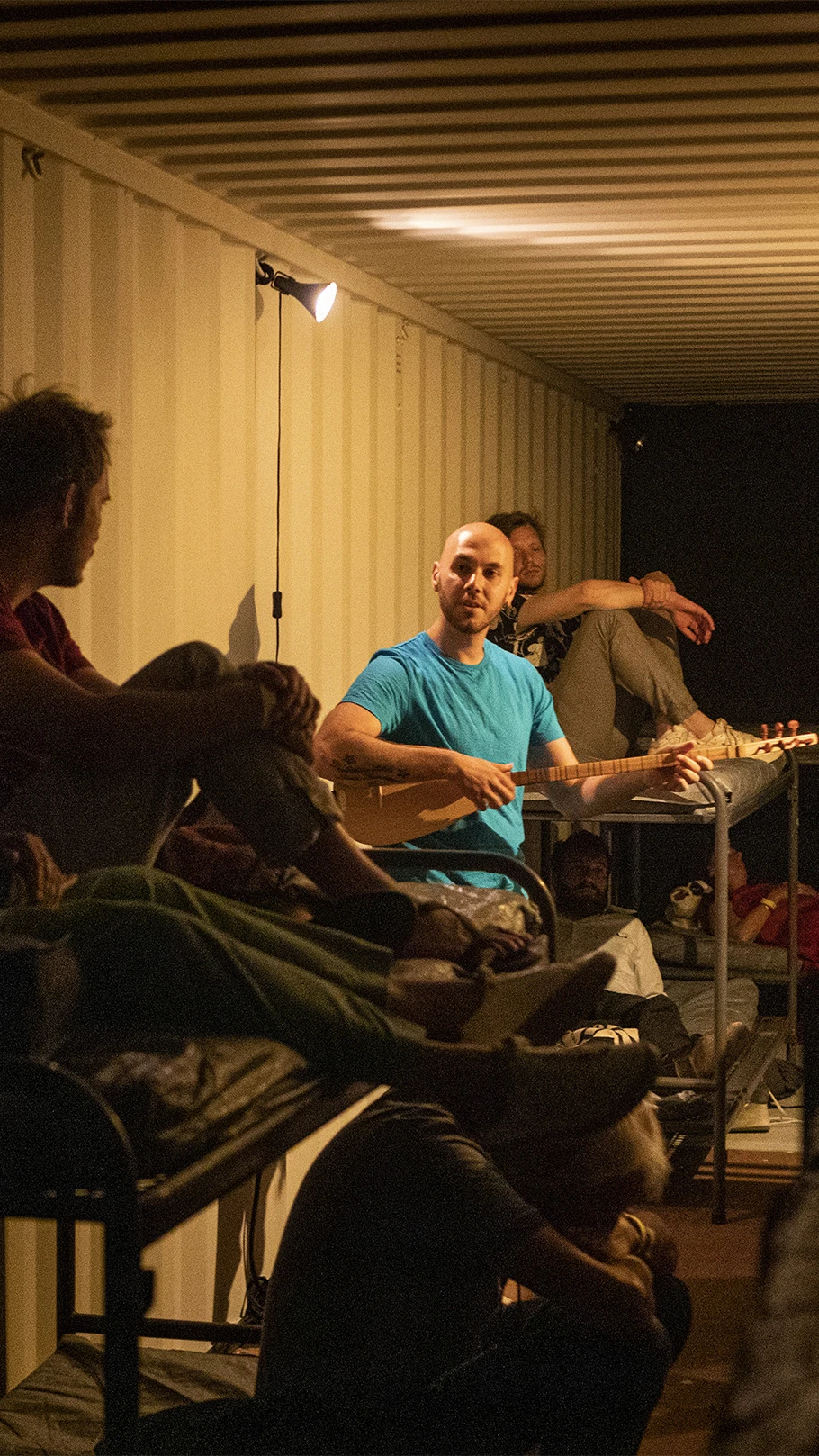 A group of people in a dimly lit shipping container, some seated on bunk beds.