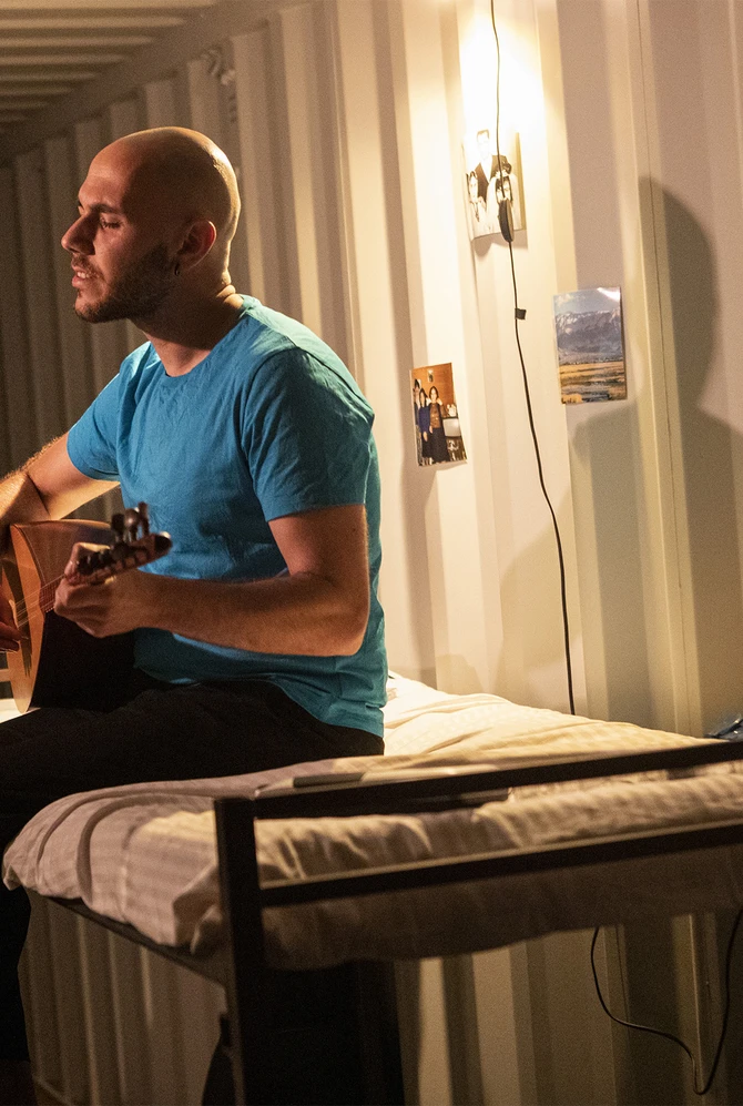 Man in a blue shirt plays a guitar while sitting on a bed in a dimly lit room.