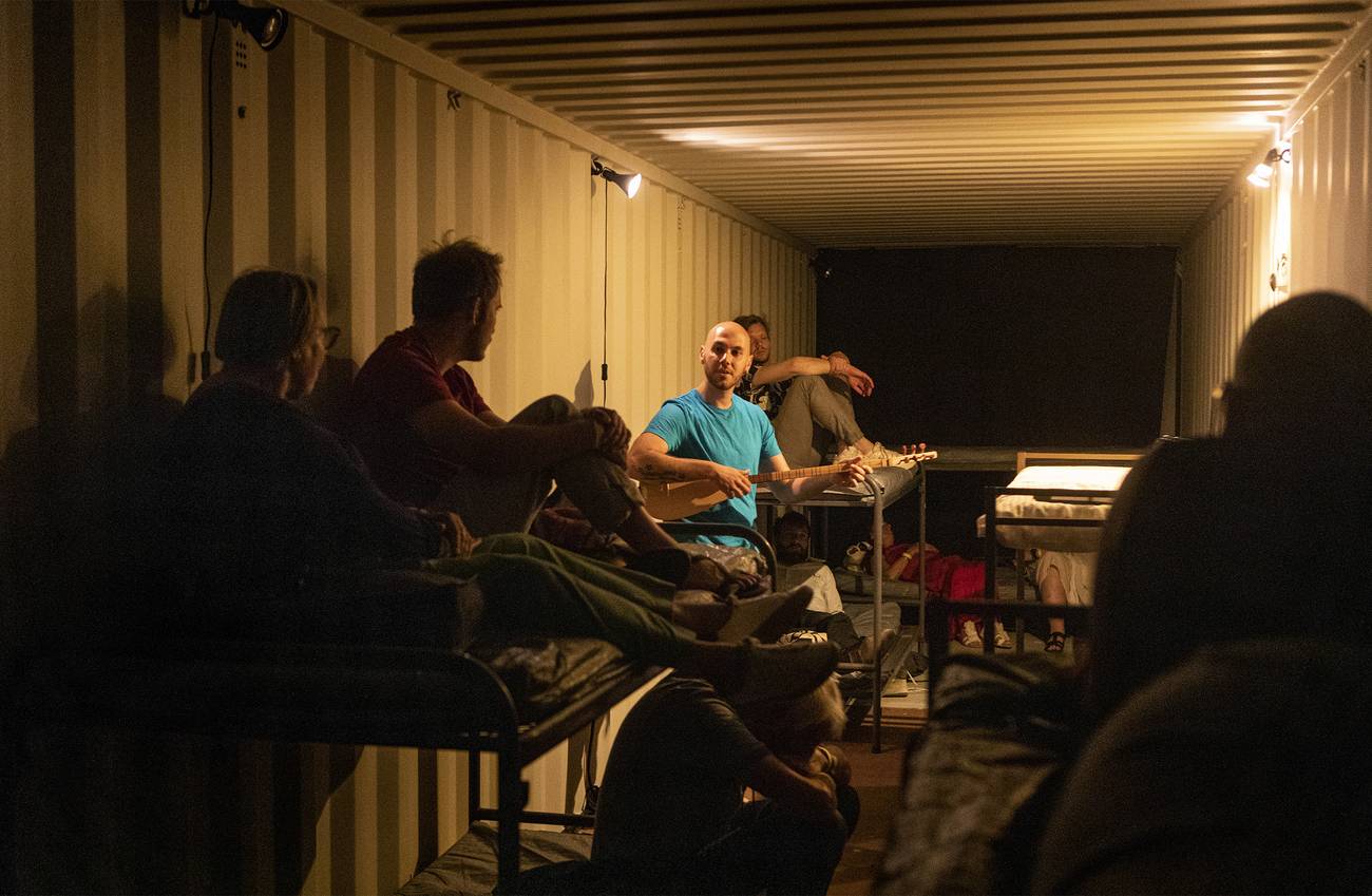 A group of people in a dimly lit shipping container, some seated on bunk beds.