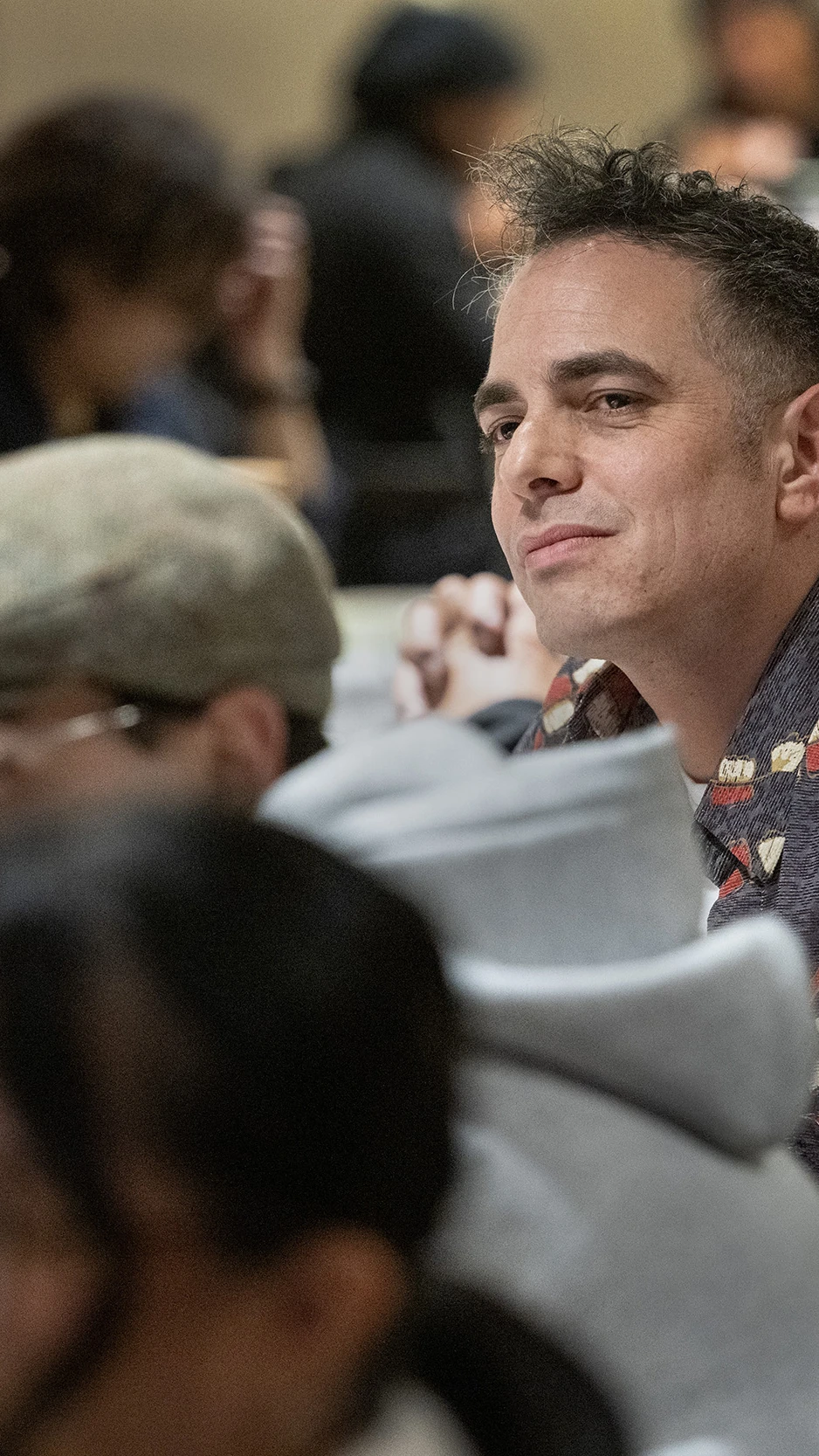A man in a patterned shirt sits at a crowded event, looking content.