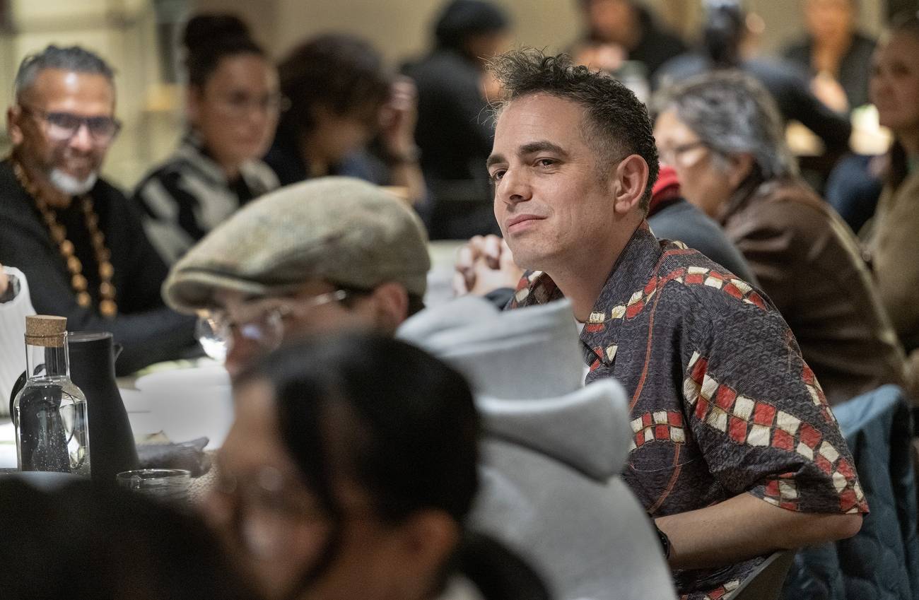 A man in a patterned shirt sits at a crowded event, looking content.