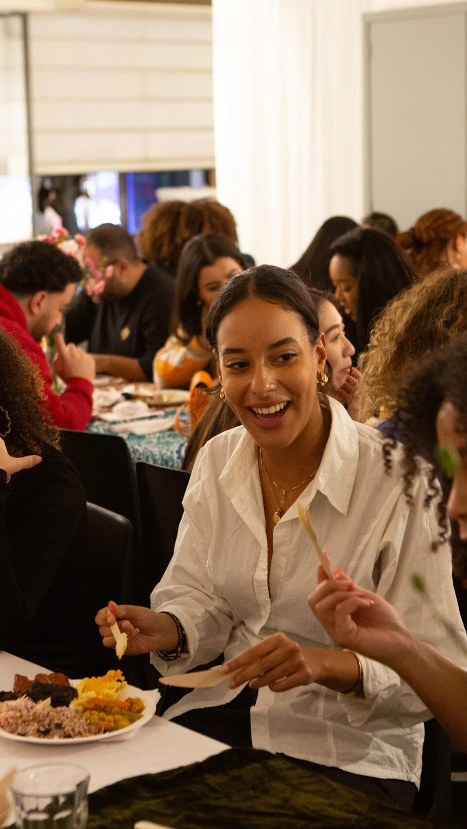 Mensen zitten aan lange tafels samen te eten. De sfeer is vrolijk en er staan bloemen op tafel.