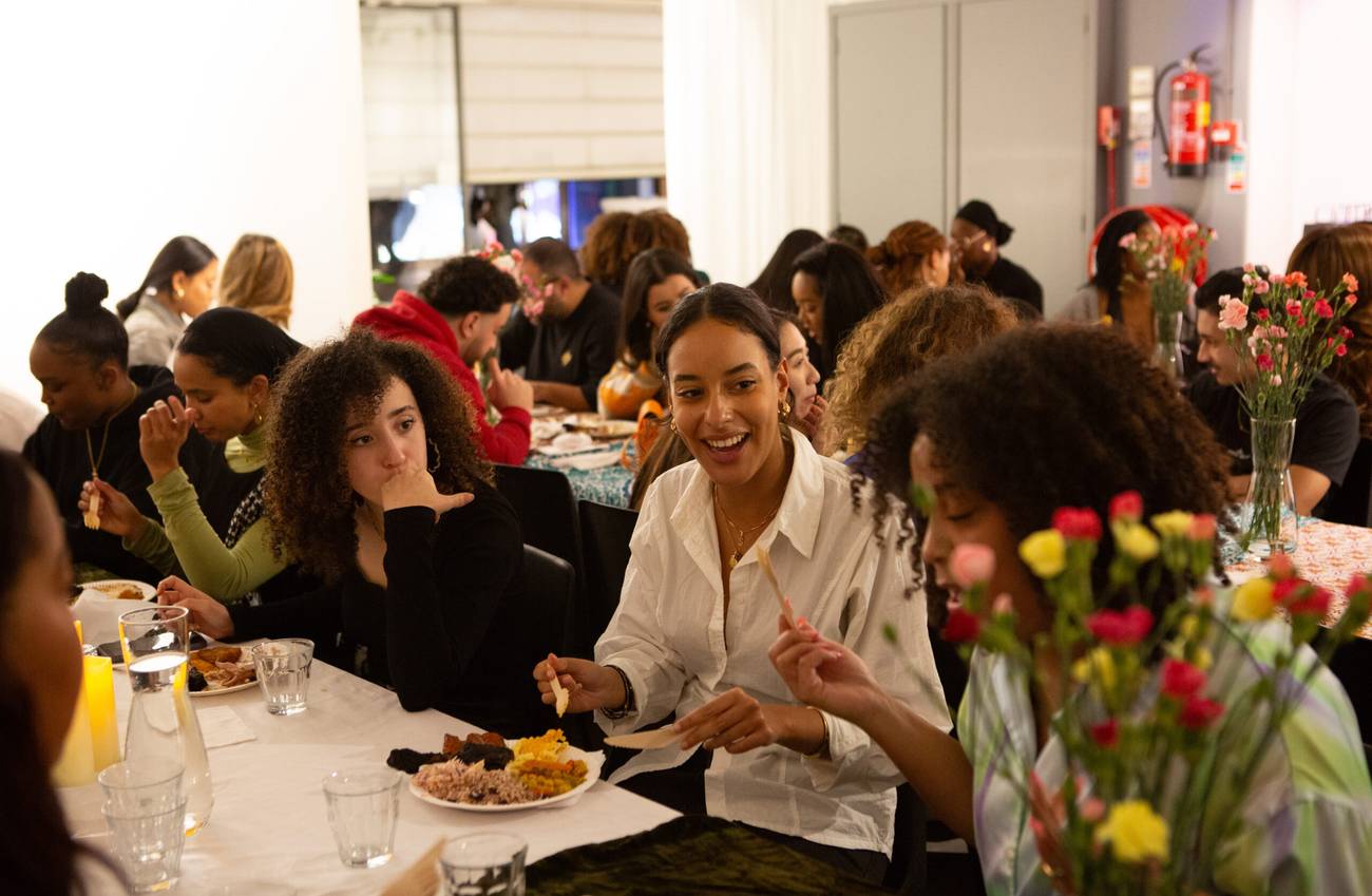 Mensen zitten aan lange tafels samen te eten. De sfeer is vrolijk en er staan bloemen op tafel.